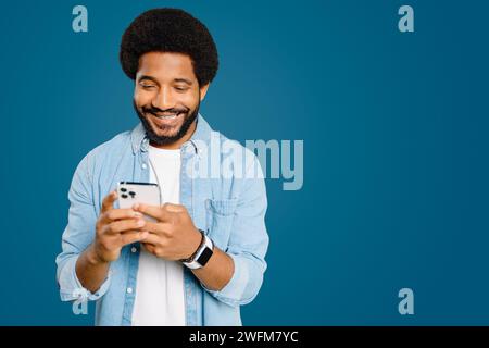 Junger lateinischer Mann mit Afro-Haarstrahlen, während er ein Smartphone benutzt und online vor blauem Hintergrund SMS schreibt. Dieses Bild repräsentiert moderne Kommunikation und die Freude, in Verbindung zu bleiben Stockfoto