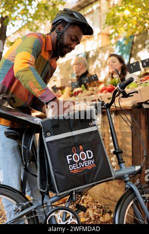 Lieferung von Frischprodukten. Junger afrikanischer Kurier, der Bestellungen mit gesunden Bio-Lebensmitteln vom lokalen Bauernmarkt abholt und selbstgewachsenes Obst und Gemüse an Kunden liefert Stockfoto