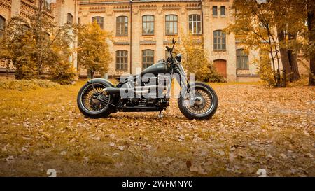 Maßgeschneidertes Motorrad basierend auf Harley-Davidson Fat Boy Modell. Seitenansicht des schwarzen Motorrads im Park in der Nähe des Gebäudes aus dem frühen 20. Jahrhundert. Stockfoto