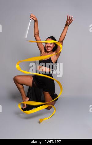Turnerin, kniend auf dem Boden, Bewegungen mit gelbem Band. Isoliert auf grauem Hintergrund. Stockfoto