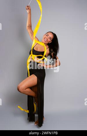 Schöne Turnerin, die akrobatische Bewegungen mit einem gelben Band macht. Isoliert auf grauem Hintergrund. Stockfoto