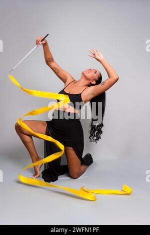 Turnerin, kniend auf dem Boden, Bewegungen mit gelbem Band. Isoliert auf grauem Hintergrund. Stockfoto