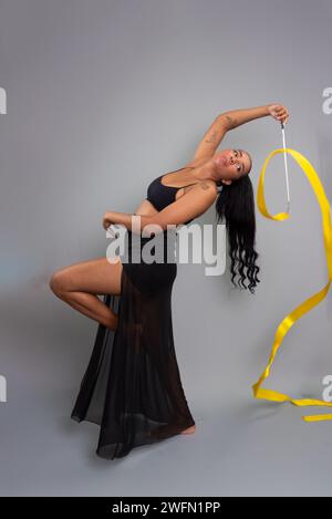 Schöne Turnerin, die akrobatische Bewegungen mit einem gelben Band hinter ihrem Rücken macht. Isoliert auf grauem Hintergrund. Stockfoto