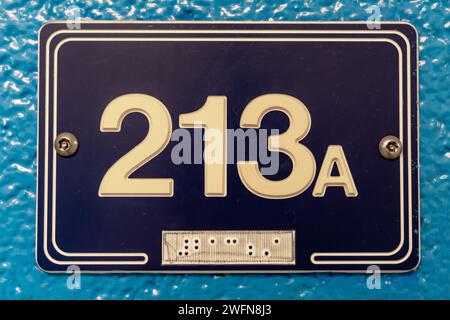 Blaues 213A-Schild mit weißem Schriftzug und braille-schrift auf blauer Blockwand. Stockfoto