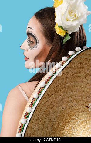 Junge Frau mit bemaltem Schädel im Gesicht und Sombrero-Hut auf hellblauem Hintergrund. Feier des mexikanischen Todestages (El Dia de Muertos) Stockfoto