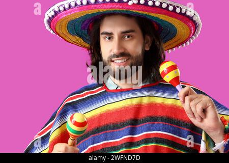 Hübscher junger mexikanischer Mann in Sombrero, Poncho und mit Maracas auf magentafarbenem Hintergrund Stockfoto