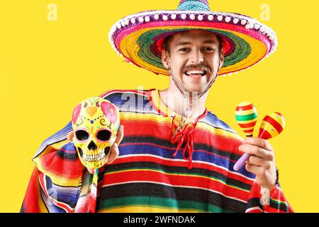 Schöner junger mexikanischer Mann mit bemaltem Schädel und Maracas auf gelbem Hintergrund. Feierlichkeiten zum Tag der Toten Stockfoto