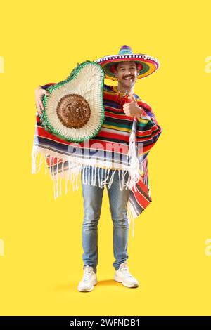 Glücklicher junger mexikanischer Mann in Sombrero, Poncho und Avocado Pinata mit Daumen nach oben auf gelbem Hintergrund Stockfoto