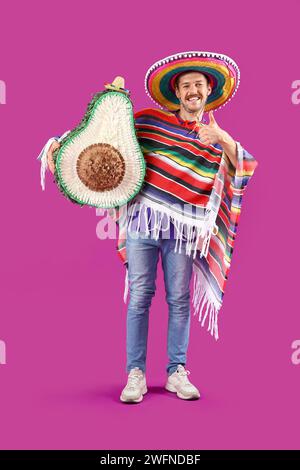 Junger mexikanischer Mann mit Avocado Pinata mit Daumen nach oben auf magentafarbenem Hintergrund Stockfoto