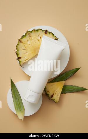 Ein weißes Röhrchen auf runden Podesten, dargestellt mit Ananasblättern und -Scheiben. Leeres Etikett zur Förderung von aus Ananas gewonnenen Schönheitsprodukten Stockfoto