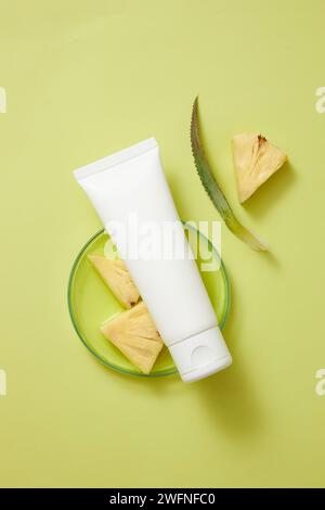 Draufsicht eines weißen Tubus mit leerem Etikett, der auf eine Petrischale aus Glas gelegt wird, die aus Ananas- und Ananasscheiben extrahiert wurde. Kosmetisches Mittel Stockfoto