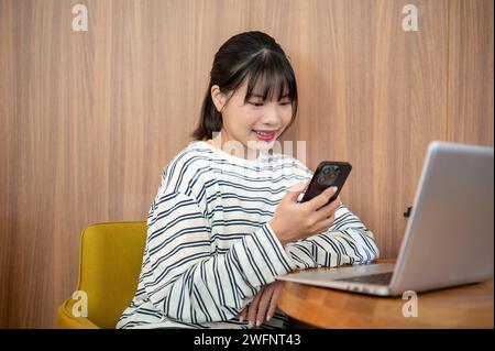 Eine positive asiatische Frau benutzt ihr Smartphone, während sie mit ihrem Laptop im Zimmer an einem Tisch sitzt, SMS liest oder Nachrichten liest. Stockfoto