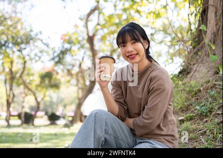 Eine entspannte und glückliche asiatische Frau in legeren Kleidern schlürft Kaffee, während sie unter einem Baum im Park sitzt und ihre Freizeit allein genießt. Menschen und LIFE Stockfoto
