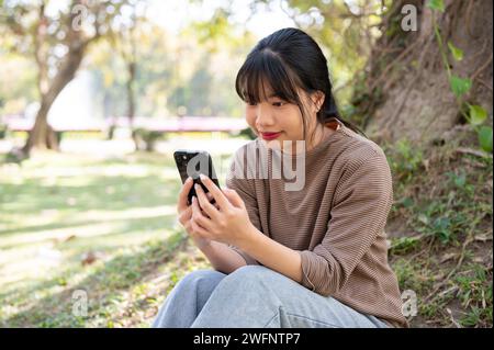 Junge asiatische Frau, die ihr Smartphone benutzt, während sie unter einem Baum im Park sitzt. Chatten, Nachrichten lesen, in sozialen Medien scrollen Stockfoto