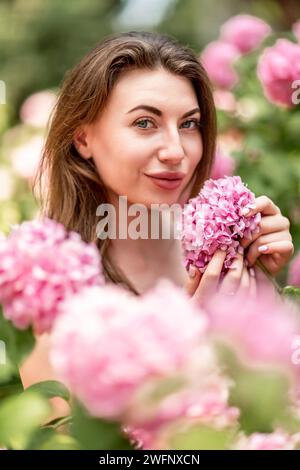 Hortensien Glückliche Frau in rosa Kleid unter Hortensien. Große rosafarbene Hortensie-Kappen umgeben die Frau. Sonniges Ambiente im Freien. Hier wird die glückliche Frau präsentiert Stockfoto