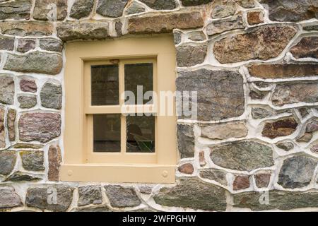Ein kleines geteiltes Fenster, das in hellbraunes Holz gerahmt ist, ist in eine Wand aus mehrfarbigem Feldstein in Valley Forge National Historical Park, Pennsylvania, eingelassen. Stockfoto