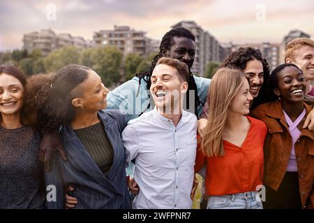 Eine überschwängliche Gruppe junger Erwachsener aus verschiedenen ethnischen Hintergründen, die einen Moment des echten Lachens und Genusses vor einer urbanen Kulisse teilen - Beau Stockfoto