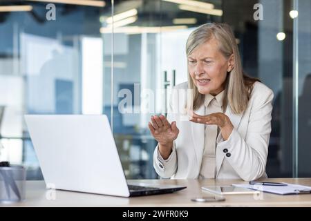 Runzelnde Seniorin, die Online-Videoanrufe per Laptop hostet und aktiv mit Händen im Glasbüro Gesten macht. Gestresste Frau, die Schwierigkeiten mit App-Verständnis und Internetverbindung hat. Stockfoto