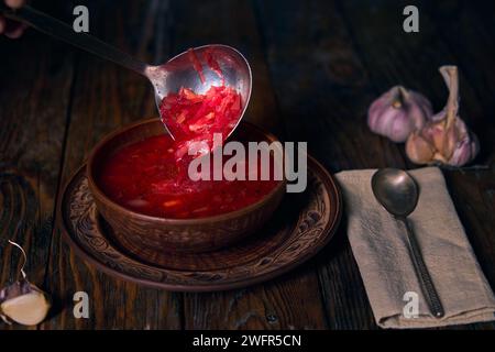 Nationaler ukrainischer Borschtsch, Steingut, Kelle, die Borschtsch in einen Teller gießt, Knoblauch, Serviette und Löffel auf einem Holztisch Stockfoto