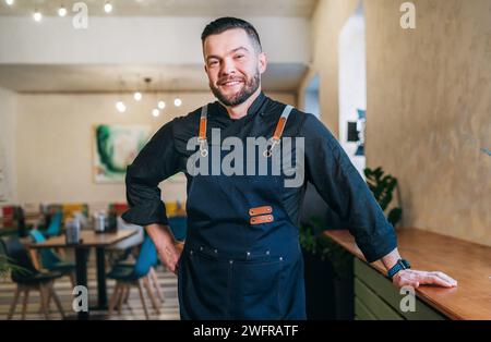 Porträt eines glücklichen, lächelnden kleinen Geschäftsinhabers in schwarzer Küchenuniform mit Schürze in seinem gemütlichen Restaurantsaal. Erfolgreiche Menschen, harte Arbeit Stockfoto