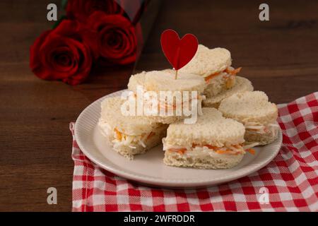 Herzförmige Sandwiches aus Salat, die auf einer weißen Schale auf einem rot-weiß karierten Tuch liegen. Auf dem Hintergrund steht ein Rosenstrauß. st. Va Stockfoto