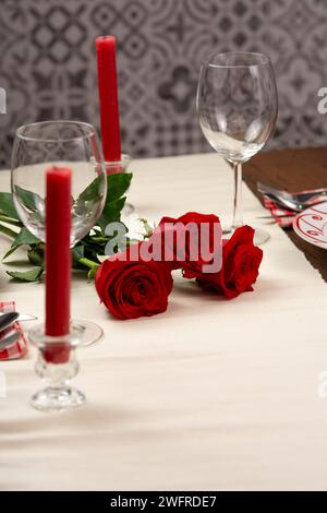 Set aus drei Rosen, die auf einer Tischdecke liegen. Auf dem Tisch stehen zwei Weingläser, zwei rote Kerzen und Besteck. Vertikales Bild Stockfoto