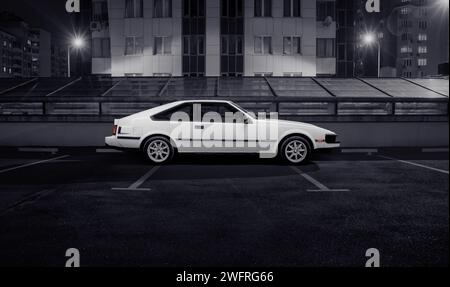 Weißes Toyota Celica Supra Liftback Coupé parkt auf der Stadtstraße. Seitenprofil eines japanischen Autos aus den 1980er Jahren vor dem nächtlichen städtischen Hintergrund. Stockfoto