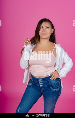 Stilvolle Frau in weißem Hemd und Jeans, die zur Seite zeigen, mit durchdachtem Ausdruck, rosa Hintergrund. Stockfoto