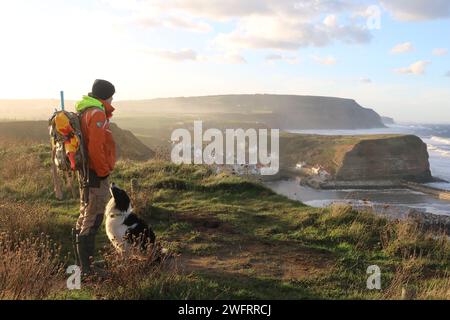 Ein Mann und sein Border Collie Hund, auf dem Cleveland Way Pfad, oberhalb des alten Fischerdorfes Staithes während eines trüben Sonnenuntergangs. Stockfoto