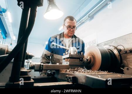 Der ältere turner, 50-55 Jahre alt, arbeitet an der Drehmaschine in der Drehwerkstatt. Vorderansicht. Authentischer Workflow. Stockfoto