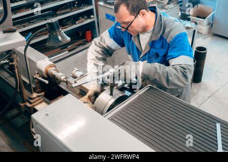 Porträt des professionellen turners bei der Arbeit an der Drehmaschine in der Werkstatt. Der 50-55-jährige turner in Overall und Brille dreht das Teil in der Werkstatt an der Maschine. Fotografie authentischer Arbeitsprozess. Stockfoto