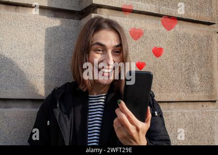 Lächelnde Frau, die ihr Smartphone ansieht, mit roten Herzen, die rauskommen Stockfoto