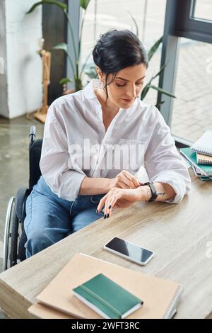 Gut aussehende engagierte behinderte Geschäftsfrau im Rollstuhl, die ihre Armbanduhr im Büro überprüft Stockfoto