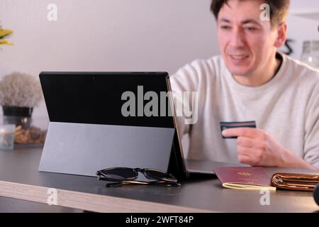 Ein glücklicher junger Mann mit Laptop und Kreditkarte in der Hand, Reisepass und Geldbörse auf dem Tisch Stockfoto