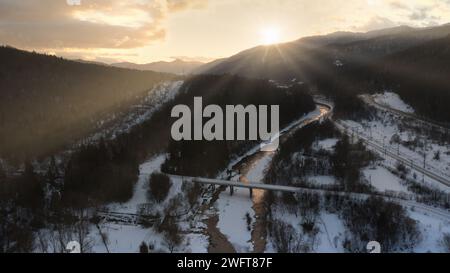 Wintersonnenaufgang über eine Brücke über einen teilweise gefrorenen Fluss mit umliegender schneebedeckter Landschaft. Malerischer Winterhintergrund. Schönheit der Natur. Stockfoto