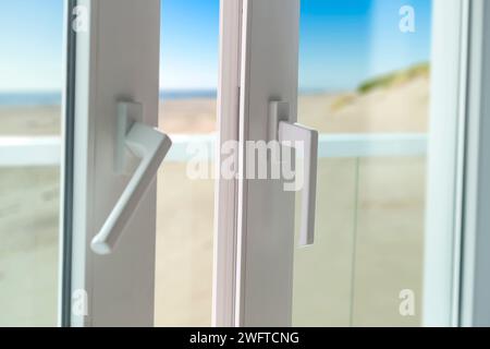Schiebetür vom Balkon zum Strand. Nahaufnahme des Türschlosses mit schöner und sonniger Meereslandschaft im Hintergrund. Weiße PVC-Tür und Sicherheitsglas. Stockfoto