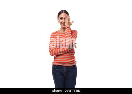 Eine 30-jährige, schlanke europäische Frau in einer lässigen orangefarbenen Bluse flirtet, während sie ihr Gesicht mit der Hand bedeckt Stockfoto