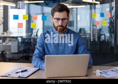 Porträt eines ernsthaften und fokussierten jungen Spezialisten, Entwicklers und Programmierers, der im Büro am Tisch sitzt und an einem Laptop arbeitet. Stockfoto