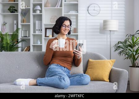 Lächelnde junge Frau, die zu Hause auf dem Sofa sitzt und sich ausruht, ihr Handy benutzt und einen Becher mit einem Drink hält. Stockfoto