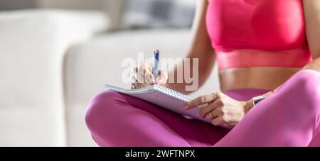 Sportliche Frau sitzt auf einer Matte und schreibt zukünftige Trainingspläne auf, um sportliche Ergebnisse zu erzielen. Stockfoto