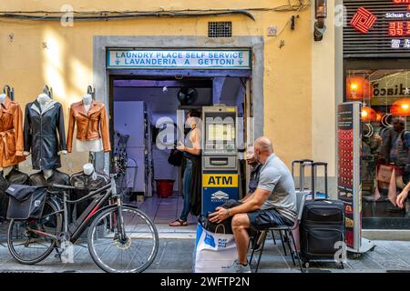 Eine Frau, die einen Waschsalon benutzt, lavanderia, gettoni, mit zwei Männern, die draußen sitzen und auf der Via Faenza warten, einer Straße in Florenz, Italien Stockfoto
