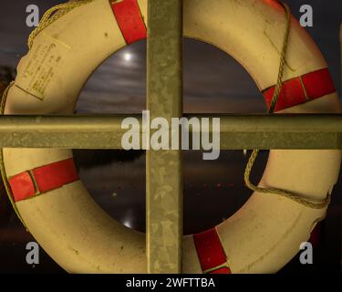 Rettungsboje, die auf dem Geländer eines Sees hängt, und der Mond reflektiert sich im Wasser. Nachteindruck. Stockfoto