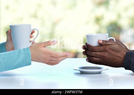 Profil mit Hintergrundbeleuchtung von Händen zweier Freunde oder Paare, die sich unterhalten und Kaffeetassen halten, die zu Hause auf einem Tisch sitzen und ein Fenster im Hintergrund haben Stockfoto
