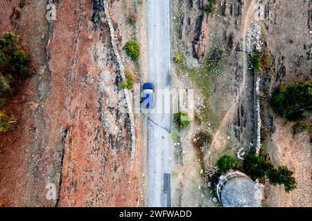 Luftdrohnenaufnahme mit blauem Auto, das auf einer schmalen Landstraße mit kargem Land an der Seite fährt, mit grünen Bäumen, die Dorfstraßen zeigen Stockfoto