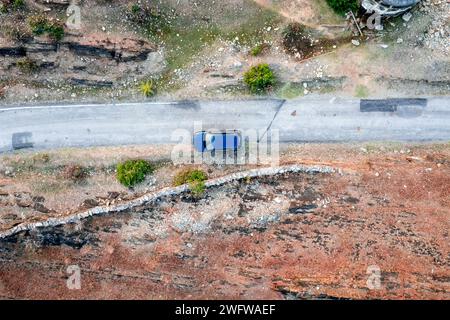 Luftdrohnenaufnahme mit blauem Auto, das auf einer schmalen Landstraße mit kargem Land an der Seite fährt, mit grünen Bäumen, die Dorfstraßen zeigen Stockfoto