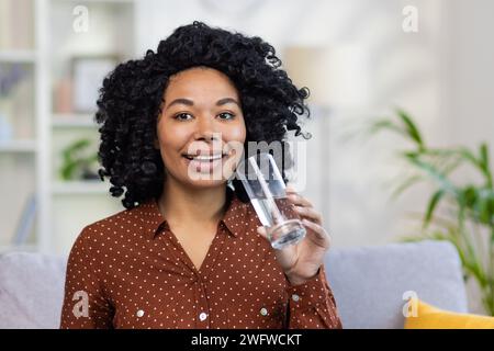 Porträt einer jungen afroamerikanischen jungen Frau, die zu Hause auf der Couch sitzt und ein Glas sauberes Wasser hält. Ich freue mich, in die Kamera zu schauen. Stockfoto