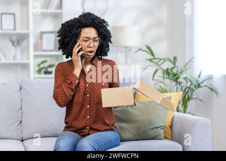Wütende afroamerikanische junge Frau, die am Telefon spricht. Er sitzt zu Hause auf dem Sofa und schaut ängstlich auf die offene Schachtel des Pakets, das er mit der Post erhalten hat. Stockfoto