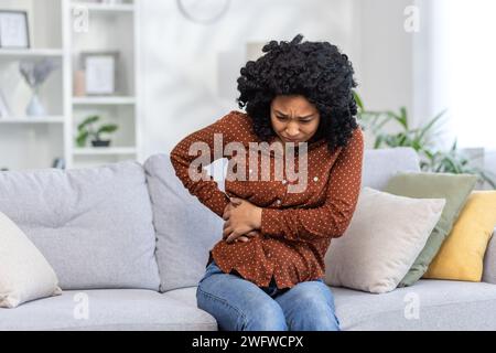 Afroamerikanische junge Frau, die zu Hause auf dem Sofa sitzt und ihre Körperseite und ihren Bauch hält. Fühlt starke Schmerzen, braucht medizinische Hilfe. Stockfoto