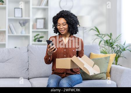 Glückliche afroamerikanische junge Frau, die zu Hause auf dem Sofa sitzt, eine offene Lieferbox hält und lächelnd auf dem Handy sitzt. Stockfoto