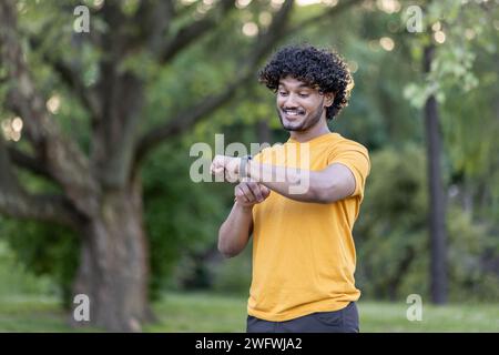 Foto eines lächelnden, modischen Indianers, der Sport treibt und im Park läuft. Steht und schaut auf die Smart Watch an seiner Hand, überprüft den Puls, erkennt die Zeit. Stockfoto